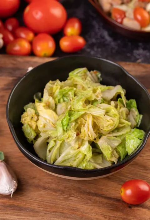 Chou sauté dans une assiette noire, accompagné d'ingrédients frais comme des tomates, de l'ail et des feuilles vertes sur un plan de travail rustique