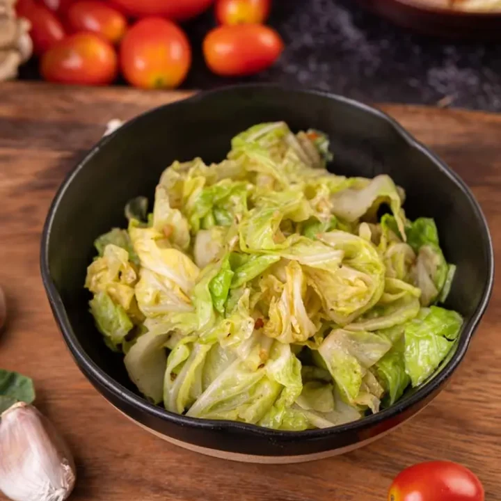 Chou sauté dans une assiette noire, accompagné d'ingrédients frais comme des tomates, de l'ail et des feuilles vertes sur un plan de travail rustique