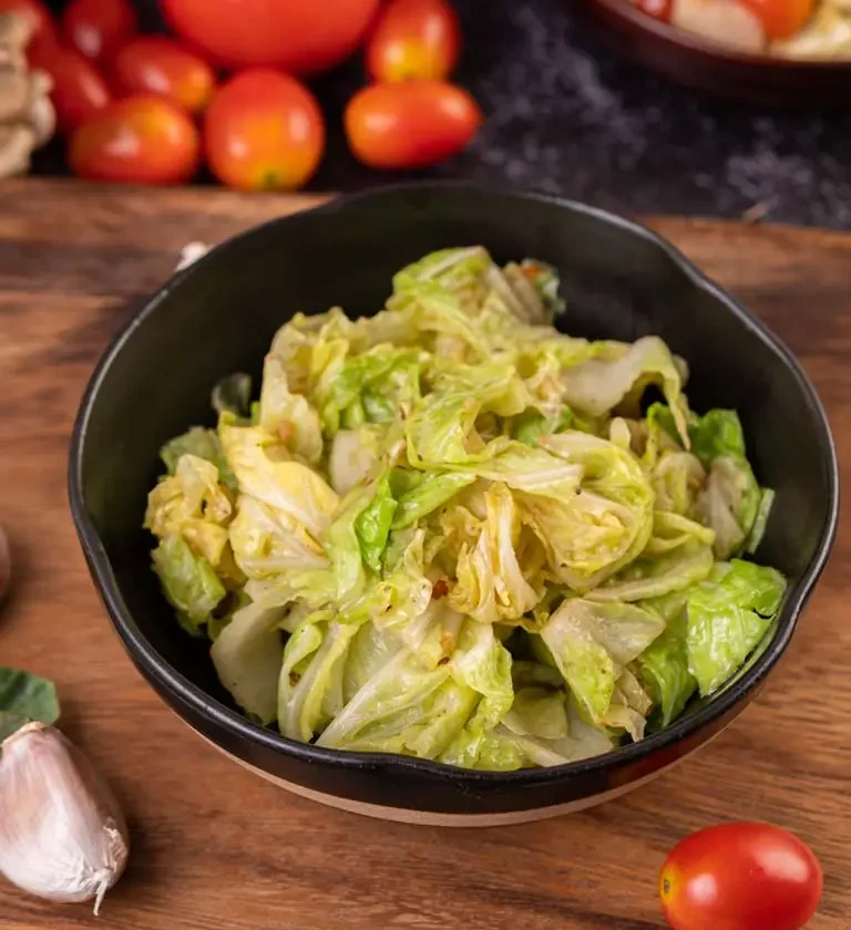 Chou sauté dans une assiette noire, accompagné d'ingrédients frais comme des tomates, de l'ail et des feuilles vertes sur un plan de travail rustique