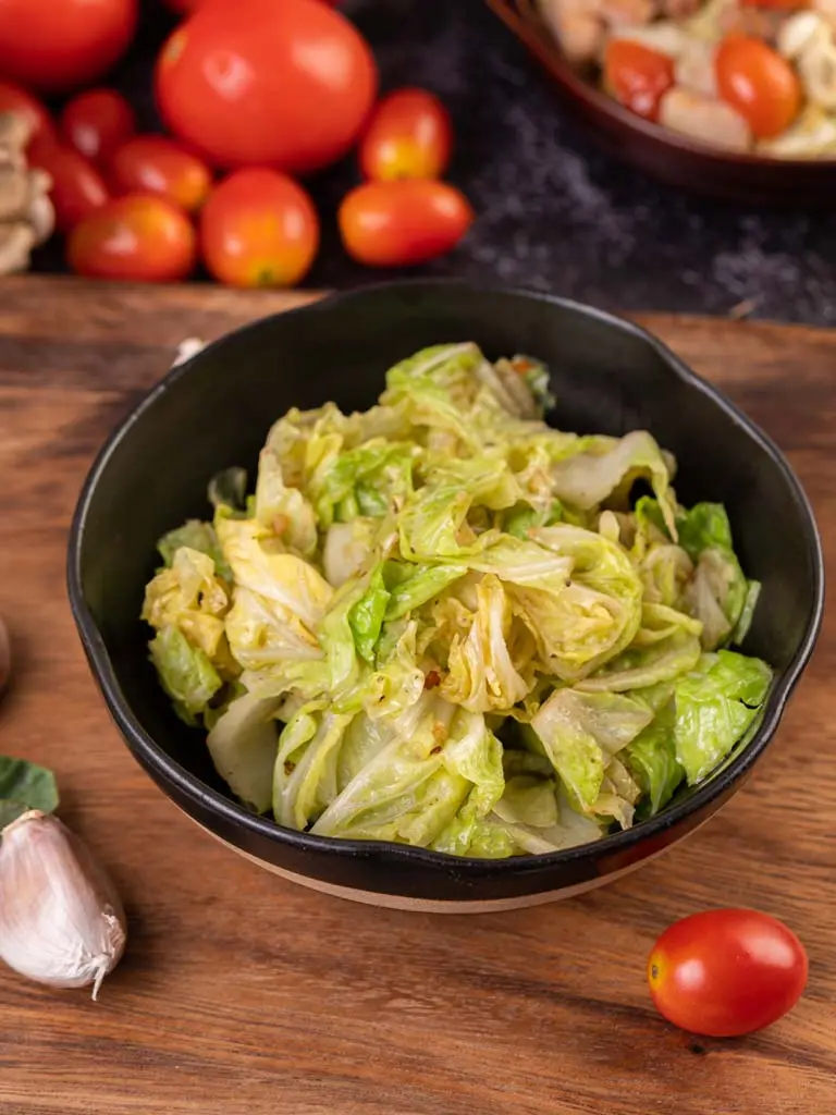 Chou sauté dans une assiette noire, accompagné d'ingrédients frais comme des tomates, de l'ail et des feuilles vertes sur un plan de travail rustique